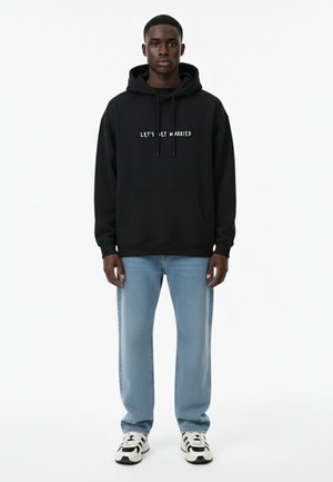 Schwarzer Hoodie mit dem Text "LASS UNS HEIRATEN", Pullover-Design, Kängurutasche, kombiniert mit hellblauen Jeans und schwarz-weißen Sneakers.