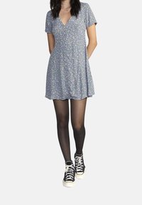 Kurzes blaues Blumenkleid mit V-Ausschnitt und kurzen Ärmeln. Kombiniert mit schwarzen Strumpfhosen und schwarzen Turnschuhen mit weißen Sohlen und Schnürsenkeln.