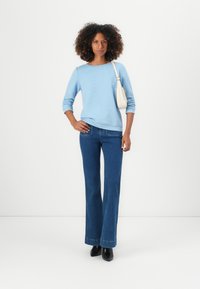 Haut long à manches longues texturé bleu clair associé à un jean évasé bleu foncé et des bottines noires. Un petit sac à bandoulière blanc complète la tenue.