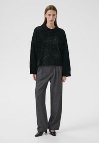 Czarny, strukturalny sweter o luźnym kroju i szerokich rękawach, w zestawieniu z szarymi, plisowanymi spodniami i czarnymi, szpiczastymi butami. Proste tło.