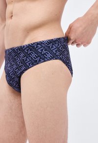 Mandlig torso iført mørke badebukser med diagonalt "EA"-mønster, hånd der justerer linningen på højre side mod en ensfarvet baggrund.