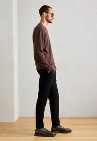 Hombre vestido con una camisa marrón de manga larga, pantalones negros, mocasines marrón oscuro y gafas de sol, de pie con las manos en los bolsillos sobre un suelo de madera clara.