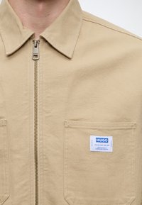 Veste beige zippée avec col, poche poitrine et étiquette blanche indiquant "HUGO" sur le coin supérieur droit de la poche.