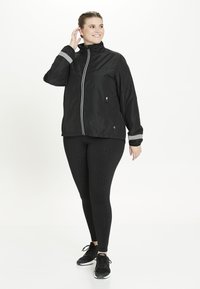 Svart lättviktsjacka med hög krage och tvåfärgad reflexdetaljer, kombinerad med svarta mönstrade leggings och atletiska sneakers.