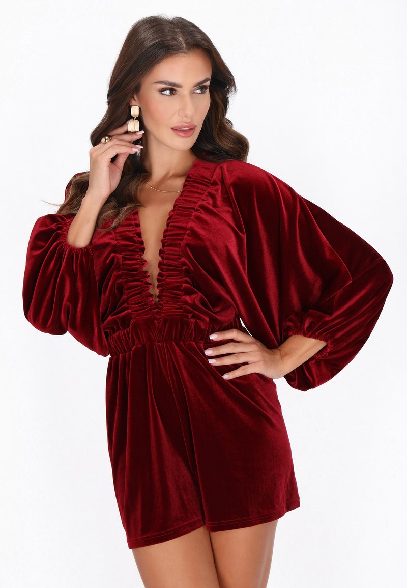 Vestido de terciopelo rojo intenso con escote pronunciado y fruncido en la parte delantera. Cuenta con mangas largas abullonadas y cintura elástica. Textura suave.
