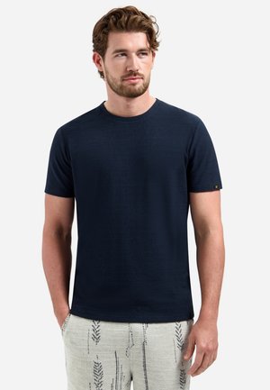 Man met kort bruin haar en baard, draagt een effen donkerblauw T-shirt met korte mouwen en lichtgekleurde broek met patroon, handen in de zakken.