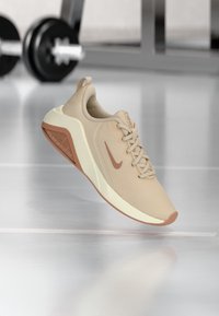 Zapatilla deportiva beige con parte superior de malla, ligero acolchado y una suela de goma marrón. Presenta un logo en el lateral y un diseño elegante y moderno.