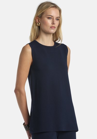 Femme blonde portant un haut bleu marine sans manches, des boucles d'oreilles en spirale dorées et un bracelet sombre, se tenant devant un fond blanc.