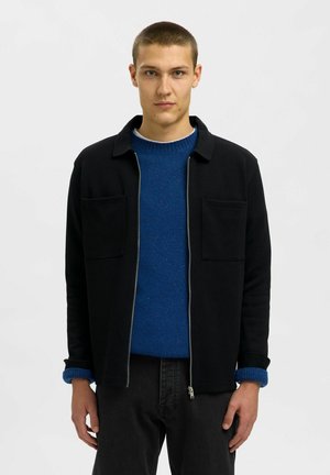 Chaqueta negra con cremallera y dos bolsillos frontales. Lleva puesta encima de un suéter azul de punto texturizado. El modelo también usa pantalones oscuros y un puño azul.