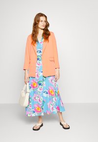 Blazer de coral sobre um vestido midi floral azul com padrões em rosa e amarelo. Uma mala de ombro bege e sandálias pretas baixas completam o visual.