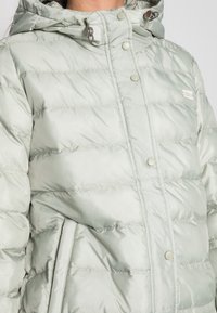 Chaqueta acolchada en verde claro con un acabado brillante. Presenta una capucha, botones de presión, bolsillos laterales y un sutil patrón de costura.