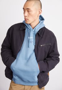 Navy jacket with a zippered chest pocket layered over a light blue hoodie. Hoodie features a printed logo and drawstring.