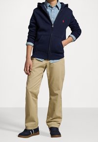 Marineblå zip-up hoodie i fleece, med framfickor och en röd logotyp. Kombinerad med khakifärgade byxor och marineblå sneakers med vita accenter.