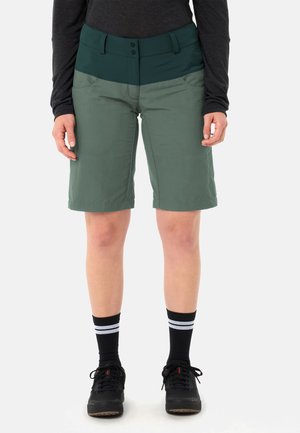 Persona che indossa pantaloncini a due tonalità di verde fino al ginocchio, maglia a maniche lunghe nera, calzini neri con strisce bianche e scarpe nere con lacci.