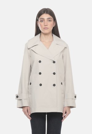 Vrouw met lang donker haar die een beige dubbelgeknoopte jas draagt met zwarte knopen en manchetriemen, staand tegen een witte achtergrond.