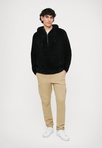 Hoodie preto felpudo com fecho na frente, combinado com calças cargo bege com bolsos laterais e ténis brancos. Design simples e cores neutras.