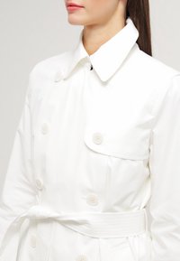 Vit trenchcoat med dubbelknäppt design, stor krage, sex knappar och bälte. Slitstark, glänsande tyg med en skräddarsydd passform.