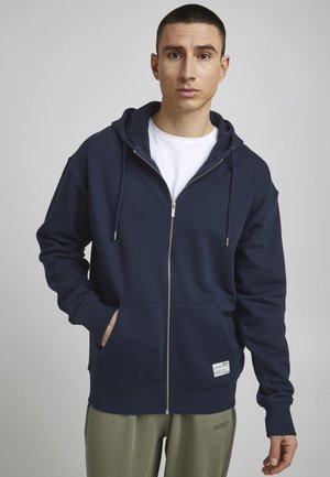 Marineblauwe zip-up hoodie van zacht materiaal, met een kangoeroezak en koordjes aan de capuchon. Gedragen over een wit T-shirt.