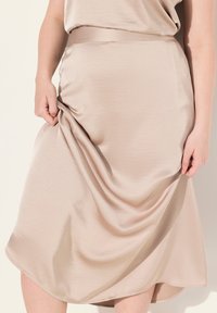 Beige midi-hame, sileä satiinikangas, näkyvä vyötärönauha, laskostettu rakenne pehmeällä kiillolla, pyöristetty helma, hienovaraisesti aaltoileva muoto.