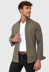 Camisa de botón en verde oliva de tejido suave, con una camiseta blanca de cuello en V debajo. Pantalones negros y cinturón marrón completan el conjunto.