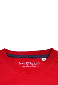Punainen puuvillapaidan t-paita, jossa on pyöreä kaula-aukko. Musta sisäkaulus ja valkoinen "Band of Rascals" -logoteksti kaula-aukossa. Kokomerkin näkyvissä.