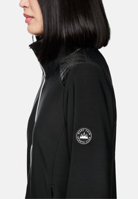 Schwarze Zip-Jacke mit gesteppten Schulterakzenten und einem runden Logo am Ärmel. Glatter Stoff und taillierte Passform, mit seitlichem Reißverschlussfach.