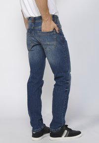 Jeans de mezclilla azul oscuro con un diseño ajustado, bolsillos traseros visibles y un ligero desgastado, combinados con zapatillas deportivas negras.