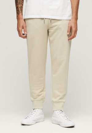Pantaloni della tuta in cotone beige con polsini a coste, vita con cordino e tasche laterali, indossati con sneakers bianche.