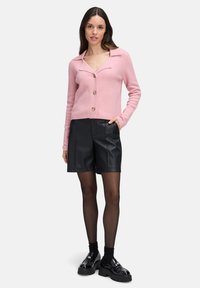 Roze wolmix cardigan met een notch-kraag en bruine knopen, gecombineerd met zwarte nepleer shorts en stevige zwarte schoenen.