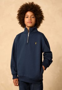 Navy pullover sweatshirt with a zip collar, ribbed cuffs, and hem. Features a small yellow emblem on the chest. Soft fabric texture.