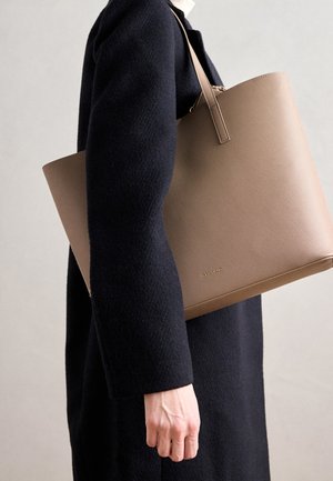 Personne en manteau sombre portant un grand sac cabas beige avec des bretelles sur le bras, sur un fond neutre.