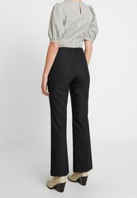 Pantalon noir évasé avec une texture lisse, taille haute et coupe ajustée. Porté avec un haut à manches bouffantes à carreaux et des bottines à talons de couleur claire.