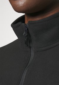 Gros plan d'une personne portant une veste polaire noire avec un col montant et une fermeture éclair noire partiellement ouverte.