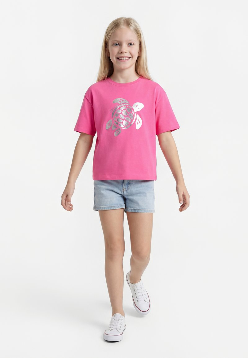 Fiatal lány sétál, rózsaszín pólót visel ezüst teknős grafikával, világos farmer rövidnadrágot és fehér tornacipőt egyszerű fehér háttér előtt.