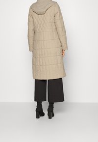 Abrigo largo acolchado beige con capucha, con patrón de rombos y bolsillos laterales, combinado con pantalones negros de pierna ancha y botas de tacón.