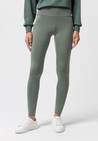 Oliwkowe legginsy z wysokim stanem, wykonane z elastycznego materiału. Mają gładką fakturę i dopasowany krój, w połączeniu z białymi sneakersami.