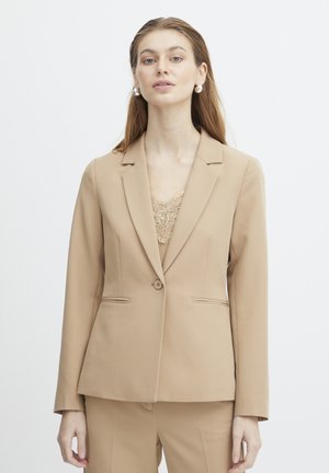 Beige bleiseri, joka on muotoiltu yksinkertaisella napilla suljettavaksi, kahdella etutaskulla ja sileällä pinnalla, käytetty pitsitopin päällä.