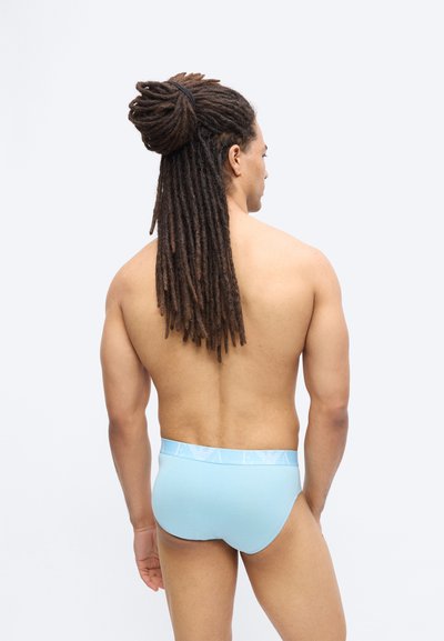 Άνδρας με μακριές κοτσίδες, δεμένες πίσω, στέκεται χωρίς πουκάμισο, φοράει ανοιχτό μπλε εσώρουχα, με την πλάτη στραμμένη σε απλό λευκό φόντο.