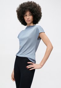 Göndör afro frizurás nő világoskék ujjatlan felsőt és fekete nadrágot viselve, kezét a csípőjén tartva fehér háttér előtt pózol.