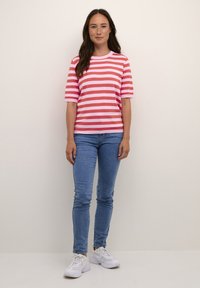 Bomullströja med rosa och ljusrosa horisontella ränder; rundad halsringning; korta ärmar. Kombinerad med tajta blå jeans och vita sneakers.
