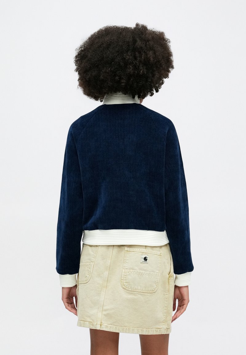 Veste en velours côtelé marine avec des poignets et un ourlet rayés blancs, associée à une jupe beige comportant une poche arrière et une étiquette.