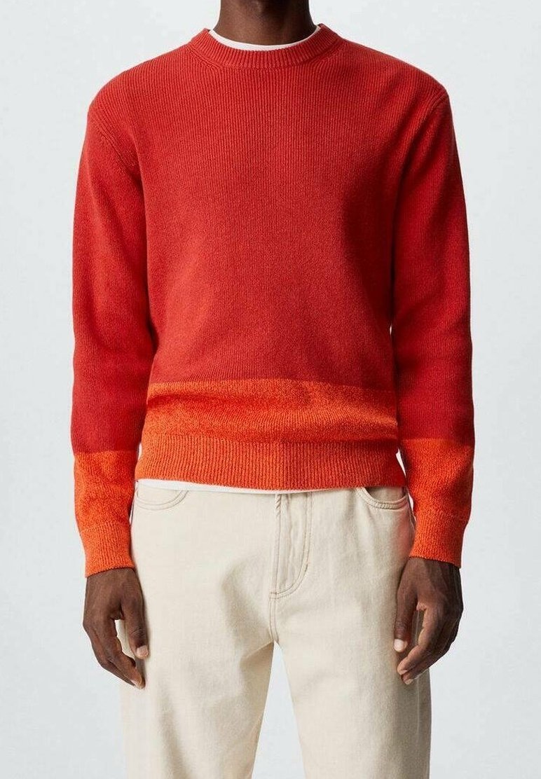 Homme portant un pull à col rond rouge-orange avec des poignets et un ourlet orange plus clair, associé à un pantalon blanc cassé sur un fond uni.