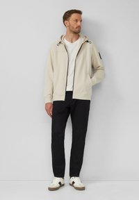 Felpa con cappuccio e zip di un beige chiaro con dettagli neri, abbinata a pantaloni scuri e sneakers bianche. Il tessuto sembra morbido con una texture liscia.