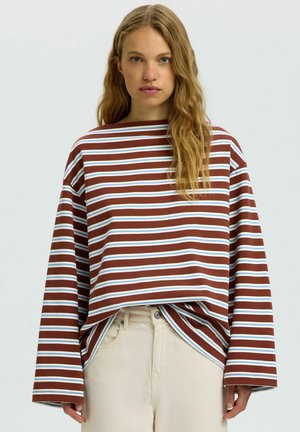 Frau mit langen, welligen Haaren, die ein braun-weiß-blau gestreiftes Langarmshirt und hellbeige Hose trägt, steht vor einem einfarbigen Hintergrund.