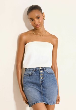 Femme portant un haut blanc bustier, des bijoux en or et une jupe en denim bleu taille haute avec boutons sur le devant, sur un fond clair uni.