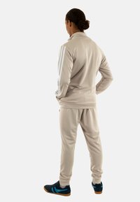 Tuta sportiva beige con giacca zip-up e pantaloni con cintura elastica, caratterizzati da tre strisce bianche sulle maniche. Scarpe nere con accenti blu.