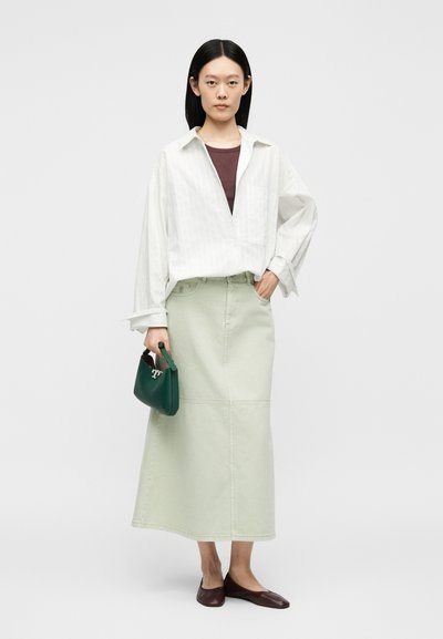 Femme debout portant une chemise blanche oversize, un top marron, une longue jupe vert clair, des ballerines marron, tenant un petit sac à main vert foncé.