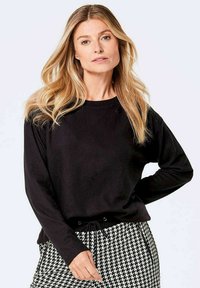 Schwarzes Langarm-Sweatshirt mit rundem Ausschnitt und verstellbarem Kordelzug am Saum, kombiniert mit einem Rock im Hahnentrittmuster.