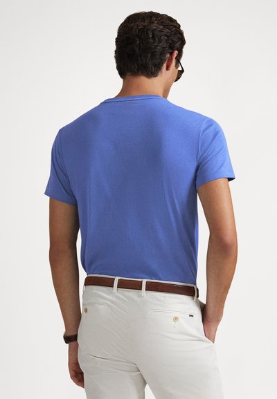 Μπλε βαμβακερό t-shirt με κοντά μανίκια, στρογγυλή λαιμόκοψη, χαλαρή εφαρμογή. Φοράει άσπρο παντελόνι και καφέ ζώνη, απεικονίζεται από την πίσω πλευρά.