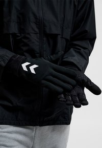 Personne portant des gants de préhension noirs avec un logo double chevron blanc, ajustant un gant sur une veste noire et un pantalon gris.
